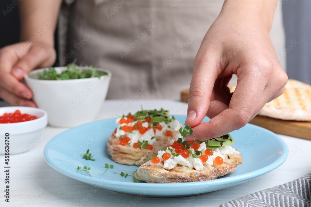 Woman putting microgreens onto sandwich with caviar at table, closeup