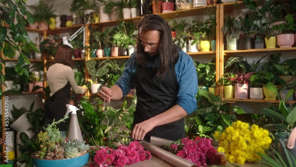 Portrait of focused professional successful male florists decorating and making bouquet with young attractive female colleague spraying and wiping plants on background. Floral concept. Portrait of focused professional successful male florists decorating and making bouquet with young attractive female colleague spraying and wiping plants on background. Floral concept.