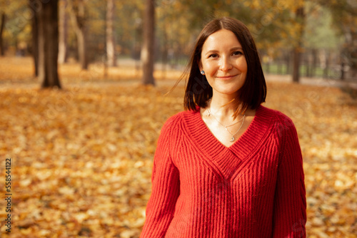 Portrait of young glad wonderful woman standing outside near trees among yellow fallen leaves in forest park, posing.