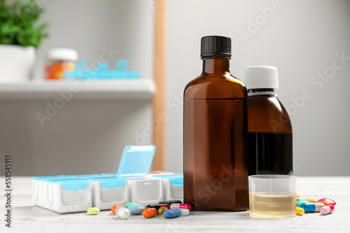Bottles of syrup, measuring cup, weekly pill organizer and pills on table. Cold medicine