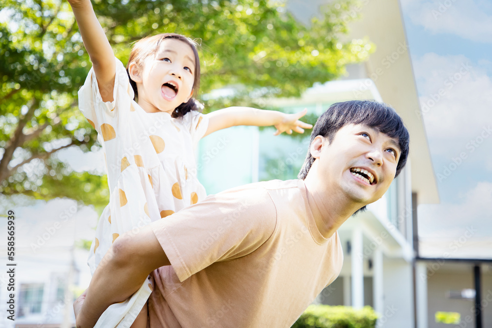 Obraz premium Attractive young father standing in garden holds on back adorable daughter at home. Smiling daughter spend time with loving father playing piggyback her daddy.