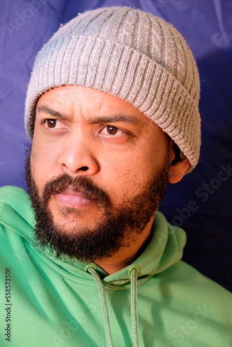 Mixed Race man with beard wearing gray beanie and green hoodie on a blue background with ring light