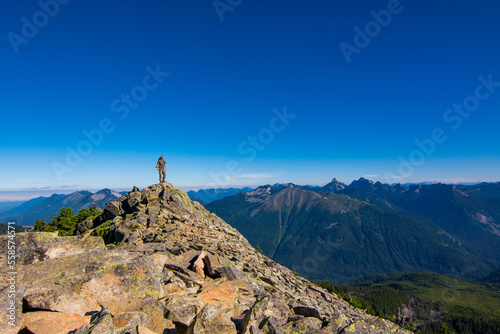 Athletic adventurous male hiker standing on top of a mountain with lush mountains in the background during a beautiful sunrise. 