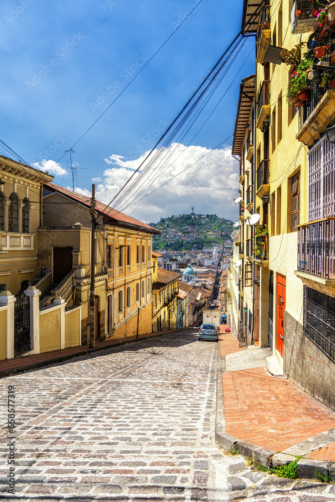 A  street in old town Quito. Colonial style buildings and. In distance hill with a statue of Virgin Mary.