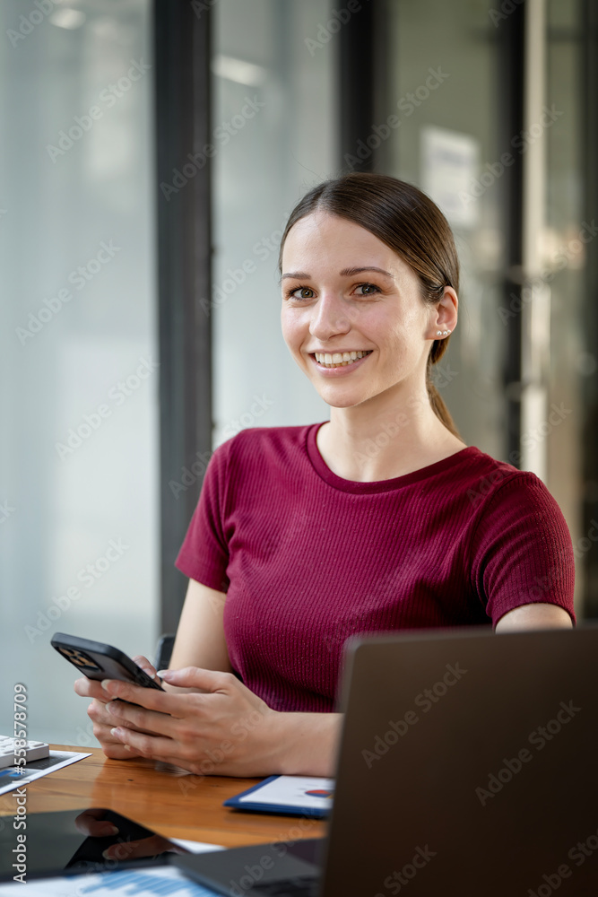 Portrait of young smiling caucasian woman holding mobile phone, sitting at office desk, looking at camera.