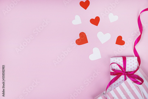Gift box with red ribbon, around lie red paper hearts. Pink background. The concept of Valentine's Day, birthday. Flat lay, top view, copy space.