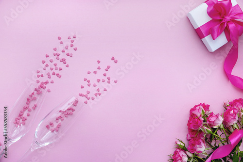 Two champagne glasses with confetti, roses and a gift box on a pink background. Flat lay, top view, copy space.