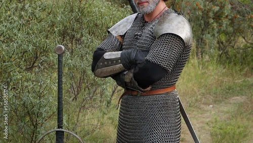 Portrait of a medieval warrior with gray hair and a scar on his face in chain mail with shoulder pads and a two-handed sword in his hands posing against the backdrop of the forest. Witcher Middle Ages