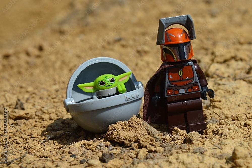 Naklejka premium LEGO Star Wars Mandalorian figure in brown suit with cloak standing on arid desert surface with alien Yoda race Child called Grogu in his flying pod, looking forward. 