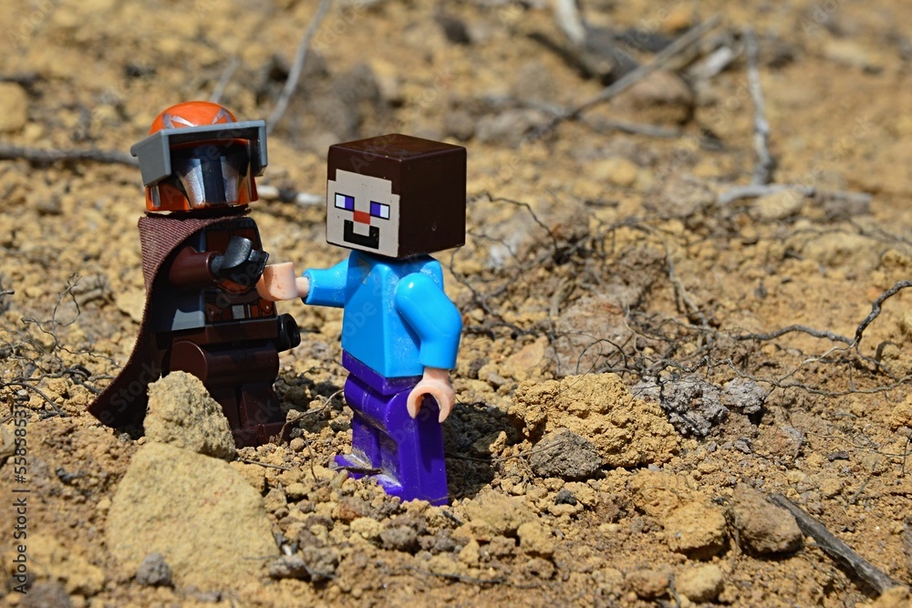 Fototapeta premium LEGO Mandalorian greets LEGO Minecraft Steve figure on arid desert like soil, summer daylight sunshine.