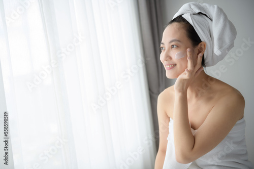 Happy young Asian woman applying face lotions while wearing a towel and touching her face. Daily makeup and skincare