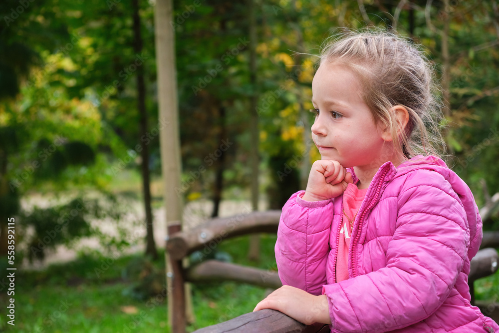 Cute children thinking outdoor park green background pink jacket hand under chin Cute children thinking outdoor park green background pink jacket hand under chin