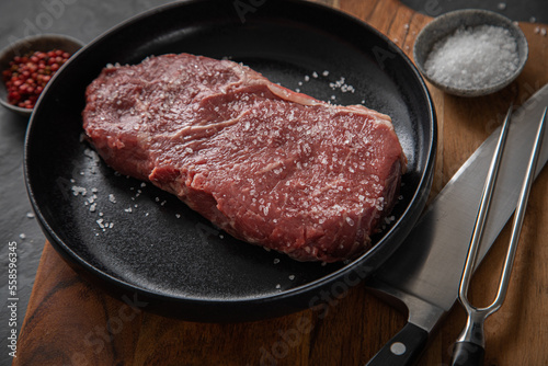 Entrecote Steak roh vom Rind mit Meersalz und roter Pfeffer auf Teller bei Vorbereitung in Küche mit Messer, Fleischgabel und Holz Brett auf Schiefer Stein Hintergrund Schwarz