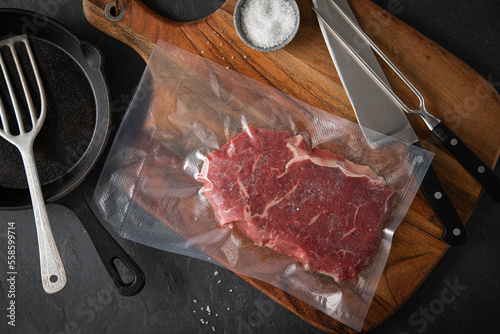 Entrecote Steak roh vakuumiert vom Rind mit Meersalz auf Teller bei Vorbereitung in Küche mit Messer, Fleischgabel, Gusseisern Pfanne, Plastik Vakuumbeutel und Pfannenwender auf Holzbrett und Schiefer