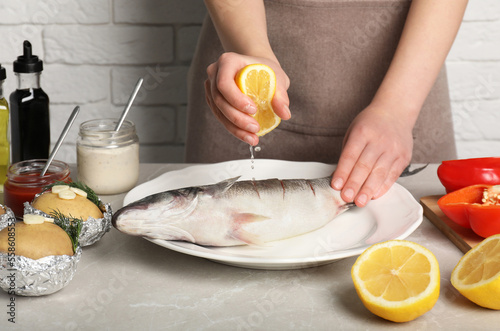 Woman squeezing lemon juice on fresh sea bass fish at light gray marble table, closeup photo