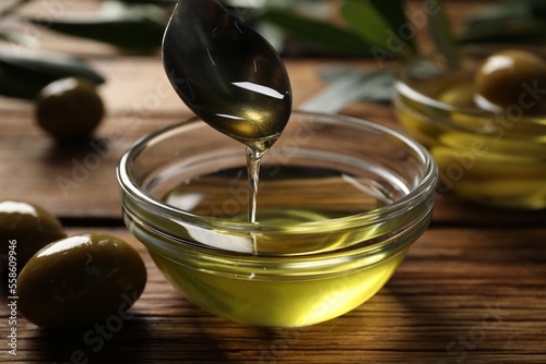 Spoon with cooking oil over bowl and olives on wooden table, closeup