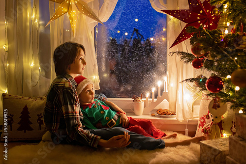 Toddler child, cute blond boy, sitting on the window in pajama, looking out for Satna Claus, Christmas lights around him © Tomsickova