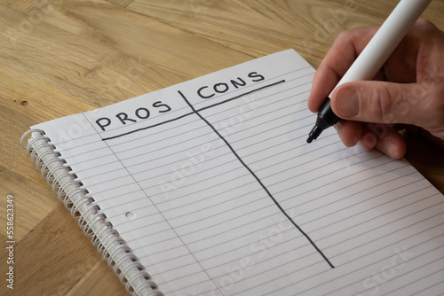 A man writes a list of pros and cons by hand in a notepad on a desk in order to attempt to foresee potential outcomes of various actions to help an important decision making process photo