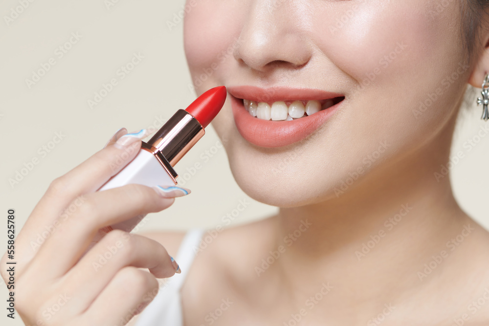 beautiful smiling asian woman holding red lipstick, isolated on beige