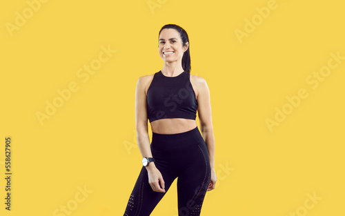 Portrait of beautiful happy successful female sports trainer isolated on yellow background. Happy millennial woman with athletic figure dressed in black in leggings and top smiling at camera. Banner.
