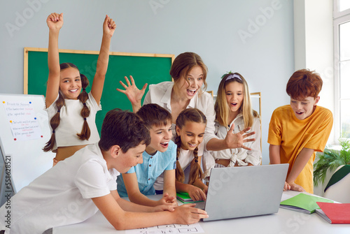 Small group of happy cheerful excited smart school children have interesting class, look at laptop computer on desk, play game, win students competition together, celebrate amazing success and victory