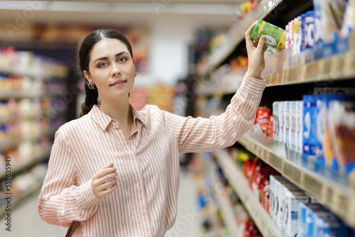 Visit grocery store. Portrait of young pretty caucasian woman takes food from upper shelve. Showcase at background. Concept of shopping and consumerism