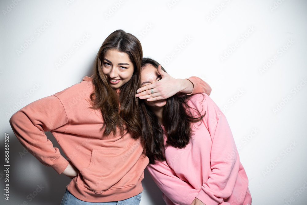 Two young attractive brunette girls in pink sweaters embracing each other smiling, one is closing the eyes of another with her hand, white background Two young attractive brunette girls in pink sweaters embracing each other smiling, one is closing the eyes of another with her hand, white background