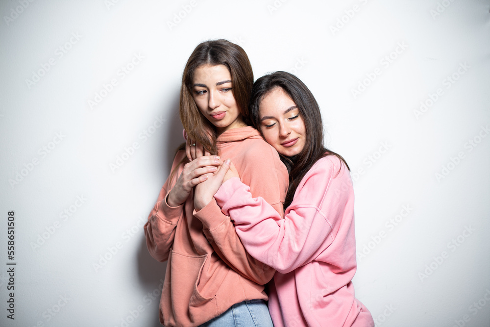 Two young attractive brunette girls in pink sweaters embracing each other, standing one after another with closed eyes, smiling, looking at the camera, white background Two young attractive brunette girls in pink sweaters embracing each other, standing one after another with closed eyes, smiling, looking at the camera, white background