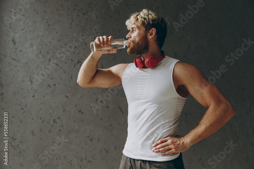 Side view young calm muscular confident strong sporty athletic sportsman man wear white tank shirt headphones drink water stan akimbo look aside warm up training indoor at gym. Workout sport concept.