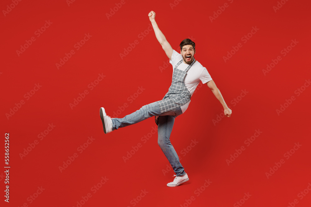 Full body side view overjoyed young male housewife housekeeper chef cook baker man wear grey apron raise up hands do winner gesture clench fists isolated on plain red background. Cooking food concept.