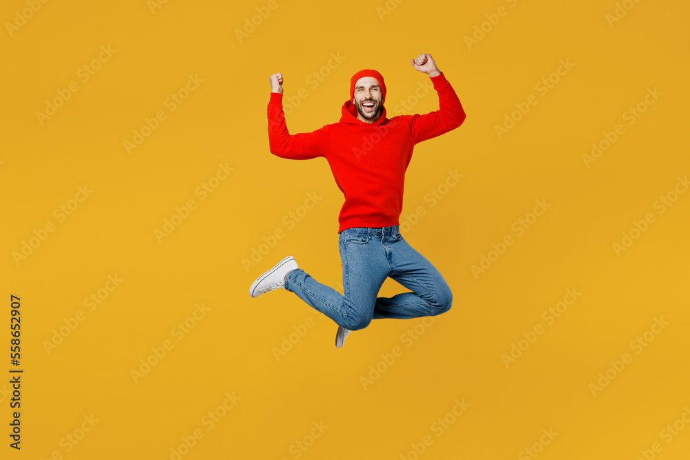 Full body young overjoyed excited fun happy caucasian man wear red hoody hat look camera jump high do winner gesture isolated on plain yellow color background studio portrait People lifestyle concept