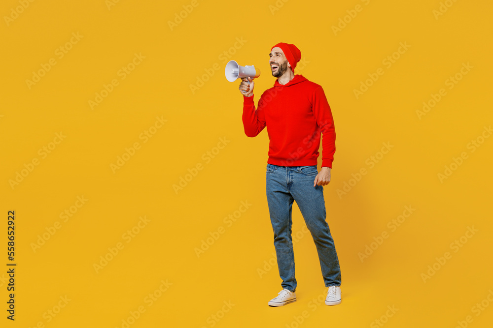 Full body young shocked excited man wears red hoody hat hold in hand megaphone scream announces discounts sale Hurry up isolated on plain yellow background studio portrait. People lifestyle concept. Full body young shocked excited man wears red hoody hat hold in hand megaphone scream announces discounts sale Hurry up isolated on plain yellow background studio portrait. People lifestyle concept.