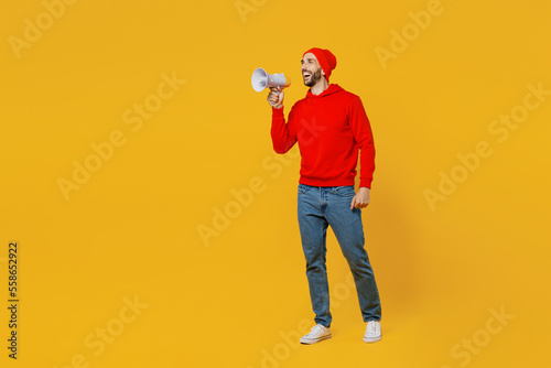 Full body young shocked excited man wears red hoody hat hold in hand megaphone scream announces discounts sale Hurry up isolated on plain yellow background studio portrait. People lifestyle concept.