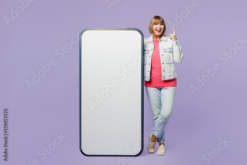 Full body elderly blonde woman 50s year old wear casual clothes denim jacket t-shirt big huge blank screen mobile cell phone with area point finger up isolated on plain pastel light purple background