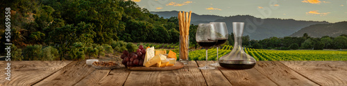 Fotótapéta Decanter and two glasses with delicious red wine, grape and cheese appetizers standing on wooden table over beautiful nature landscape of fields and forest