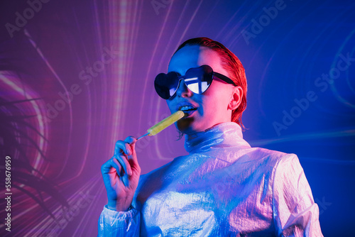 Portrait of a cyberpunk girl in heart shaped sunglasses in neon light. Seductive woman licking a lollipop in holographic clothes on the background of waves Concept of fashion, futuristic generation