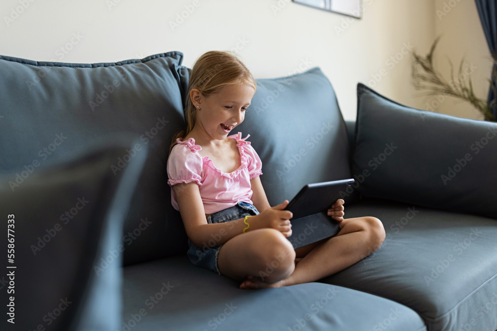 Cute little caucasian girl with blonde hair in fashionable clothes sitting at home during coronavirus pandemic quarantine and using tablet. 
