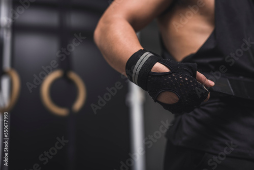 An anonymous man wearing and tank top and training gloves tightening the velcro weightlifting belt on his waist at the gym.