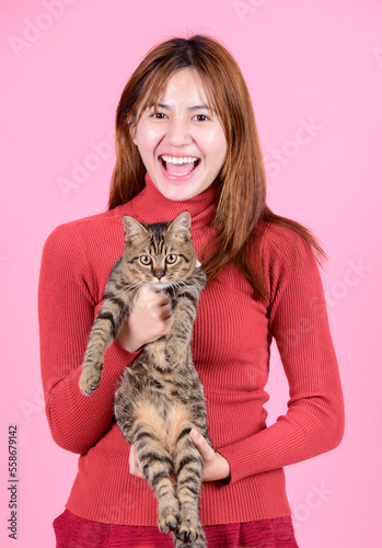 A young asian pretty cute woman in a red sweater holds a cat in her hands like a baby isolated on a bright color pink background. Asia girl with a kitten. Fiendship of pet and owner. photo