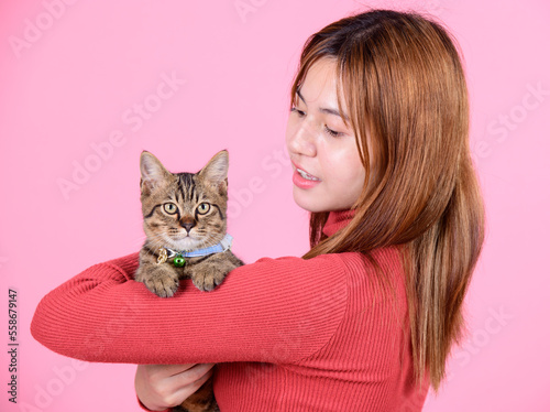 A young asian pretty cute woman in a red sweater holds a cat in her hands like a baby isolated on a bright color pink background. Asia girl with a kitten. Fiendship of pet and owner. photo