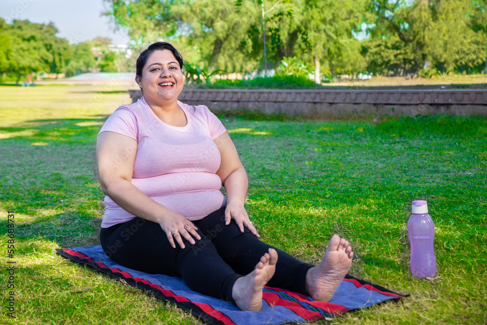 Happy Overweight Indian Woman Sitting On Yoga Mat Outdoors In Park 