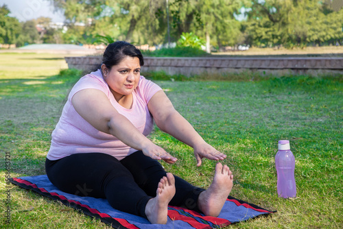 Overweight indian woman doing stretching exercise outdoors in park while watching online training classes on her laptop. Asian Fat lady Fitness lifestyle. Full length shot.