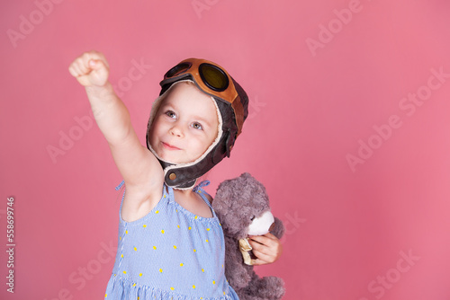 a little girl with a teddy bear in a pilot's helmet raises her hand up