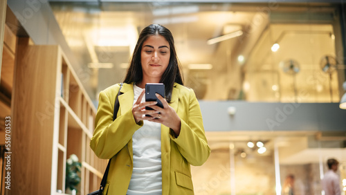 Portrait of Hispanic Businesswoman Using Smartphone App to Share Social Media Post about Career Growth. Professional Female Manager Walking Through Office Building, Smiling and Looking Happy.