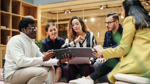 Group of People from Multiple Ethnicities Working on Problem Solving Using Notes, Laptop and Tablet in a Meeting Room at the Office. Teammates Giving Constructive Feedback on Eachother's Projects