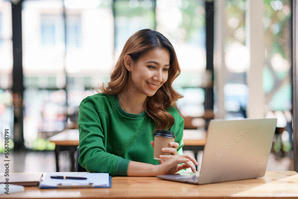 Pretty Asian businesswoman sitting happily working on her laptop and drinking coffee smiling and laughing brightly in the office. Pretty Asian businesswoman sitting happily working on her laptop and drinking coffee smiling and laughing brightly in the office.