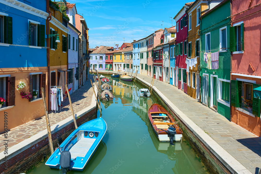 Panoramic view of canal with boats in Burano, Italy, surrounded by picturesque colorful houses decorated with plants, flowers and hanging clothes under blue sky on sunny spring day.