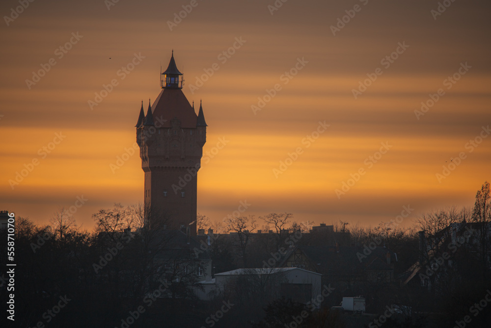 Tall tower against the orange sunset sky and surrounded by trees
