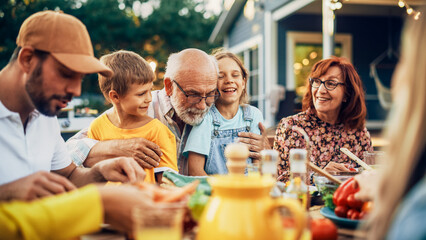 Happy Senior Grandfather Talking and Having Fun with His Grandchildren, Holding Them on Lap at a Outdoors Dinner with Food and Drinks. Adults at a Garden Party Together with Kids.