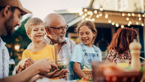 Happy Senior Grandfather Talking and Having Fun with His Grandchildren, Holding Them on Lap at a Outdoors Dinner with Food and Drinks. Adults at a Garden Party Together with Kids. photo